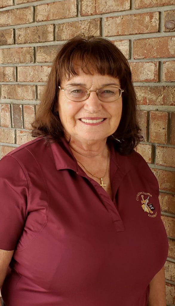 Smiling woman in a maroon polo shirt stands against a brick wall. The shirt features a logo, indicating her affiliation with a Christian home academy.