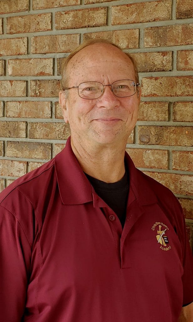 Smiling man in glasses wears a maroon polo shirt with "Christian Home Academy" logo, posed against a brick wall background.