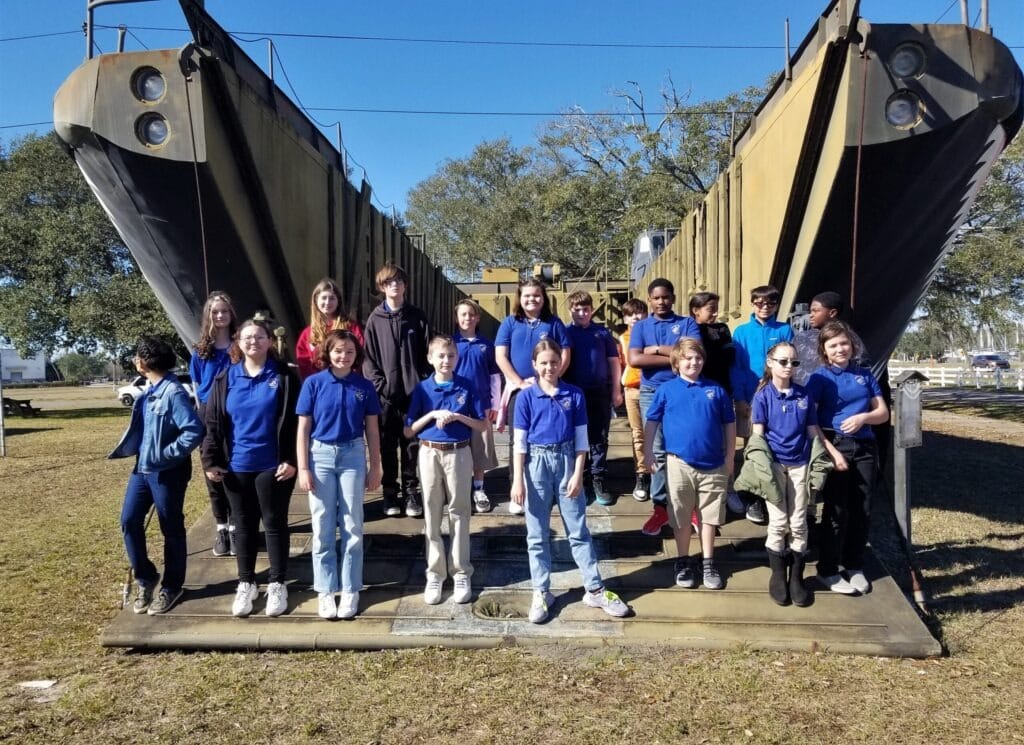 Students in blue shirts pose in front of a large military landing craft, enjoying their school field trip outdoors on a sunny day.