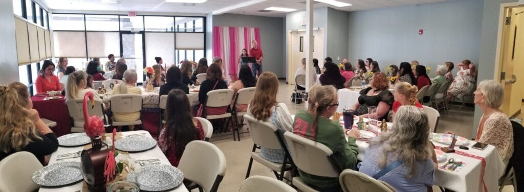 A large group of women seated at tables in a brightly lit room, listening to a speaker at a community event focused on connection and empowerment.