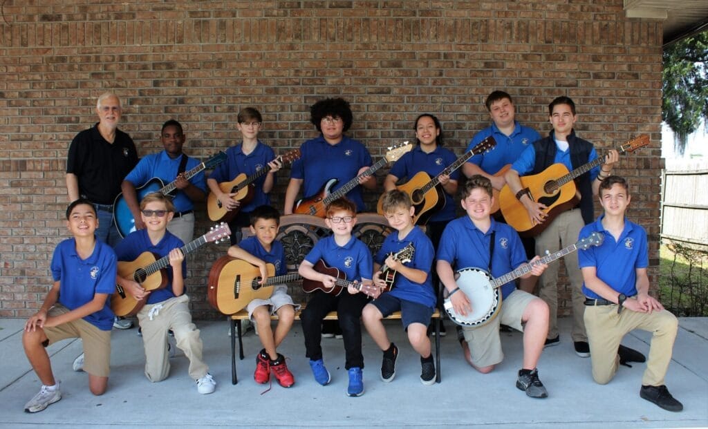 A group of twelve young musicians in blue polo shirts poses outdoors, holding guitars and a banjo, showcasing their passion for music.