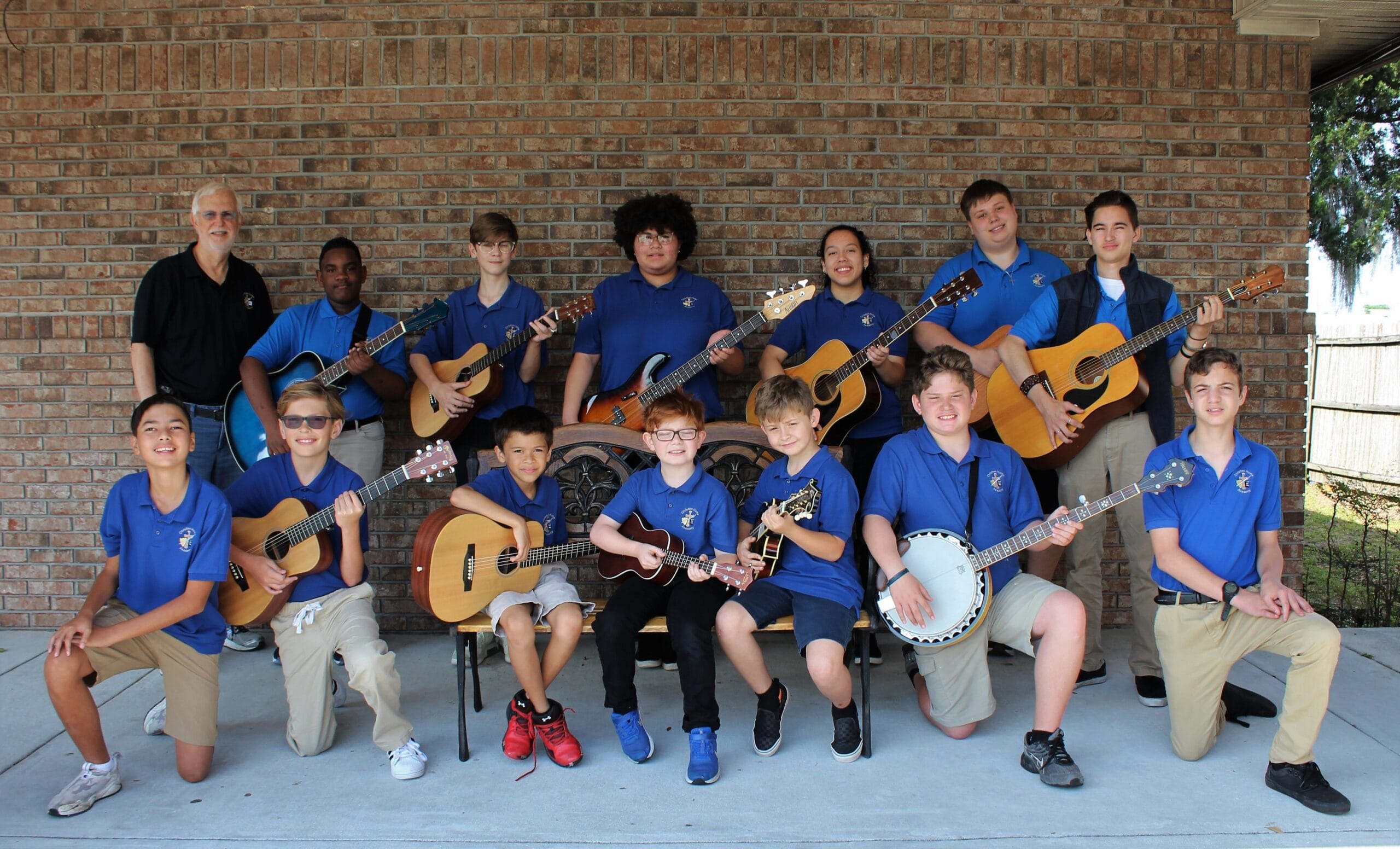 A group of twelve young musicians in blue polo shirts poses outdoors, holding guitars and a banjo, showcasing their passion for music.