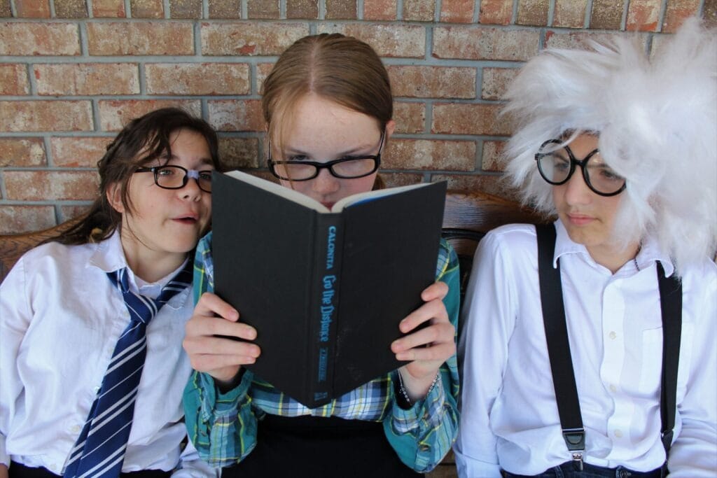 Three children dressed in quirky attire share a bench, with one reading a book titled "Go the Distance" while the others pay attention, showcasing a playful study moment.